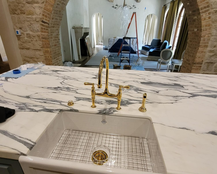 White farmhouse sink with polished brass bridge faucet on a marble kitchen island